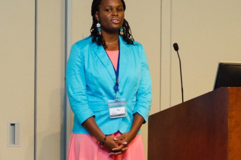 A woman stands in front of a small group speaking. To her left is a lectern with a microphone.