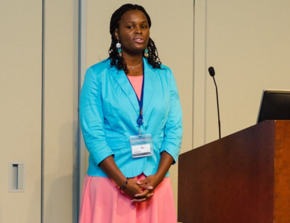 A woman stands in front of a small group speaking. To her left is a lectern with a microphone.