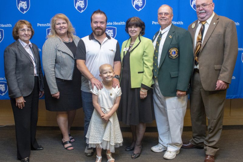 Six adults and one young girl pose for a photo. The girl is in front, smiling. The backdrop has several 'Penn State Health' logos.