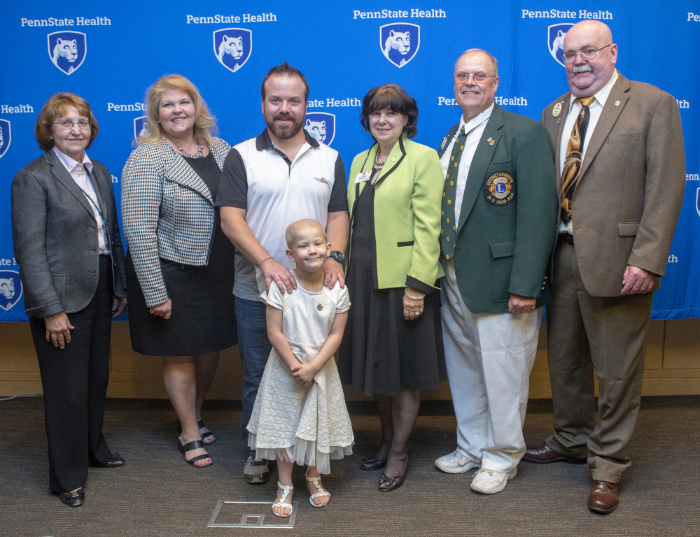 Six adults and one young girl pose for a photo. The girl is in front, smiling. The backdrop has several 'Penn State Health' logos.