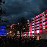 At night, the Hershey Medical Center's signature Crescent building is lit up in red, white and blue lights. Several people are sitting in the foreground.