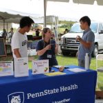 Two visiting Global Health Exchange Program students staff a table at the Farmers Market in Hershey in 2017. Three people are pictured standing behind a table in an outdoor space, with the table covered by a blue cloth with the Penn State Health logo.