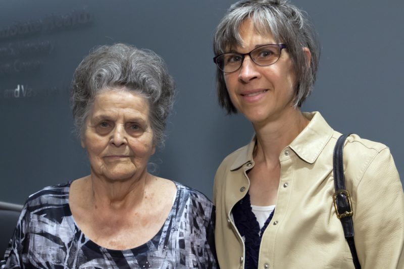 Two women stand side-by-side posing for a photo. The backdrop is a dark blue wall. A Penn State Health logo is on the wall to the left, slightly out of focus.