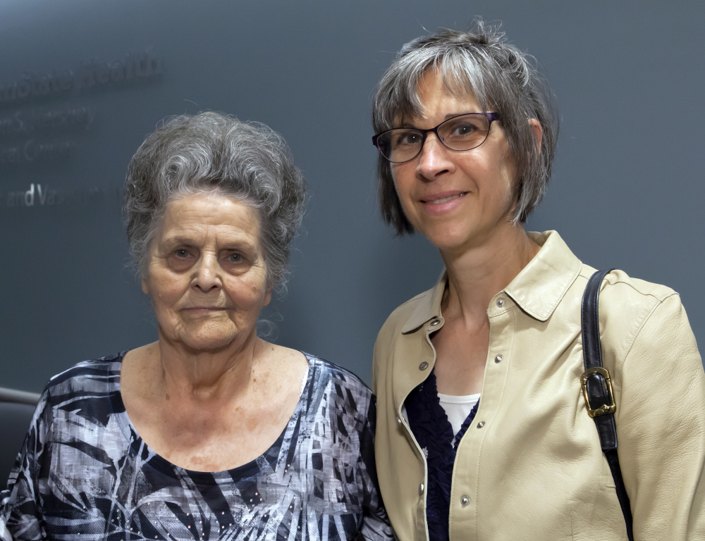 Two women stand side-by-side posing for a photo. The backdrop is a dark blue wall. A Penn State Health logo is on the wall to the left, slightly out of focus.