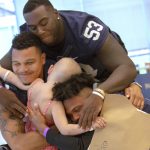 Three men in blue and white football jerseys smile as they hug a young girl. The players™ faces are facing the camera; the young girl is facing away from the camera.