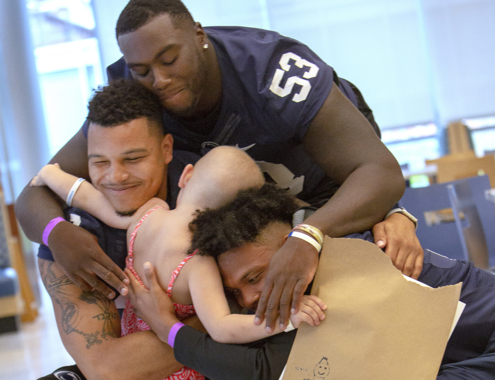 Three men in blue and white football jerseys smile as they hug a young girl. The players™ faces are facing the camera; the young girl is facing away from the camera.