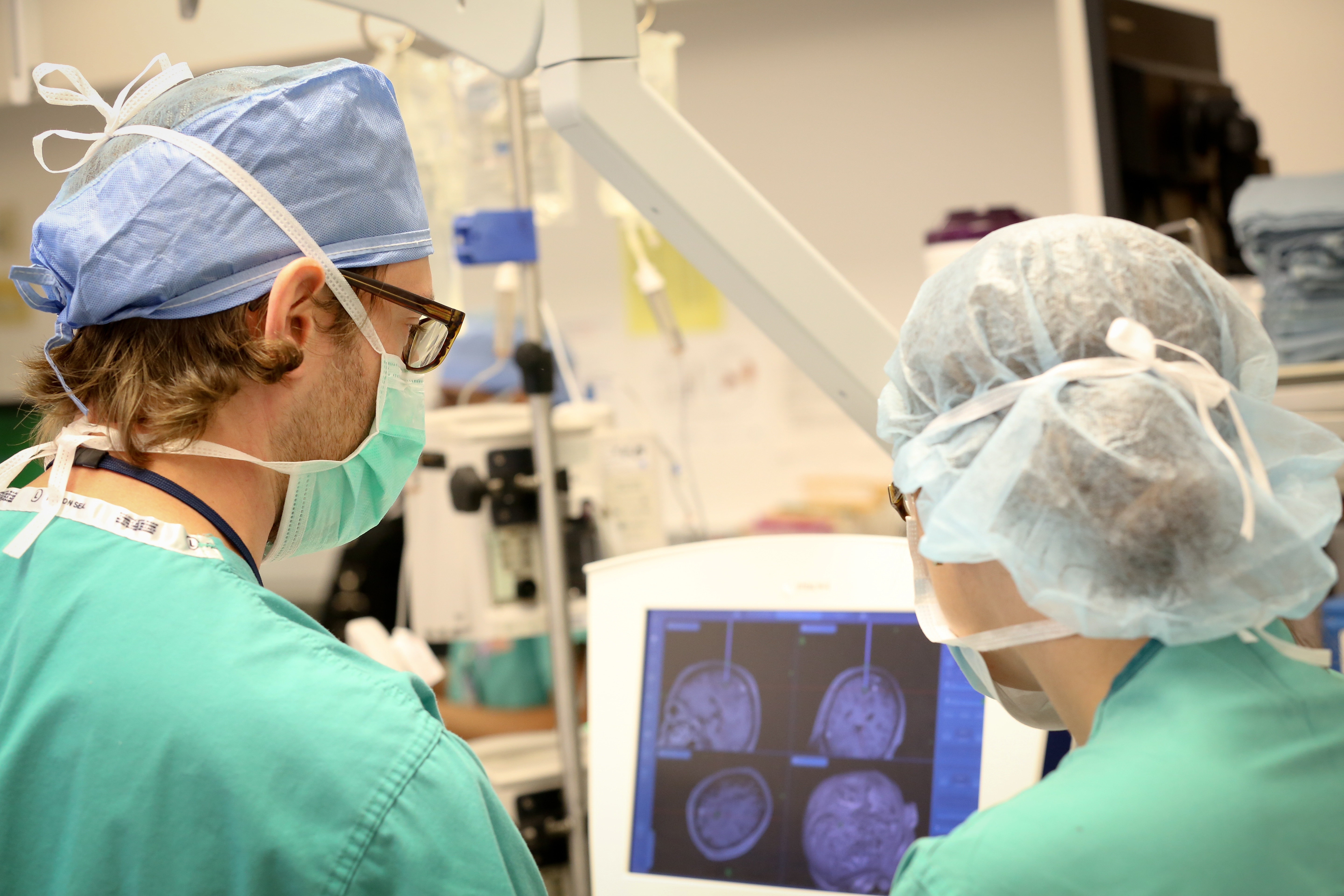 A male neurosurgeon and female neurosurgical team member look at four MRIs of a patient’s brain on a monitor. They are both wearing scrubs, surgical masks and glasses. Surgical equipment is visible in the background.