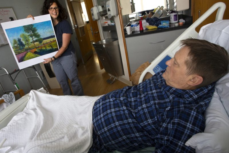 A man lays in a hospital bed. A woman stands at the foot of the bed holding a painting, showing it to him.
