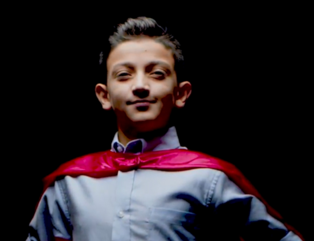 A young boy in a blue shirt and red cape looks into the camera, posing in front of a dark background.