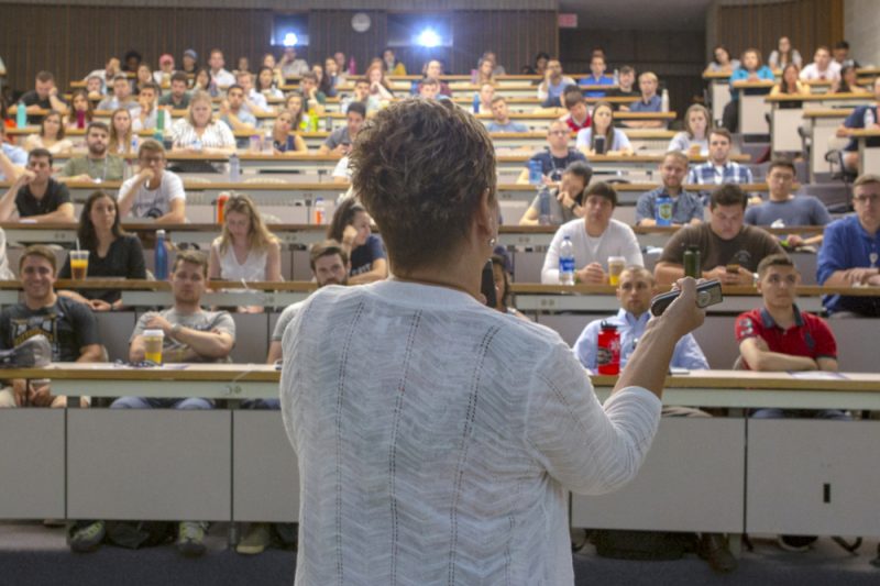 A woman in the foreground faces away from the camera, gesturing with her hands. She speaks to a crowd of people, seated in several tiered rows in a classroom.