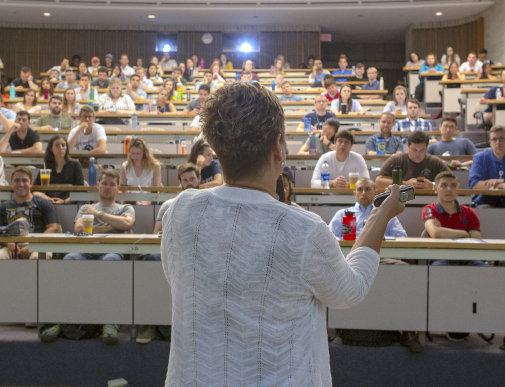 A woman in the foreground faces away from the camera, gesturing with her hands. She speaks to a crowd of people, seated in several tiered rows in a classroom.