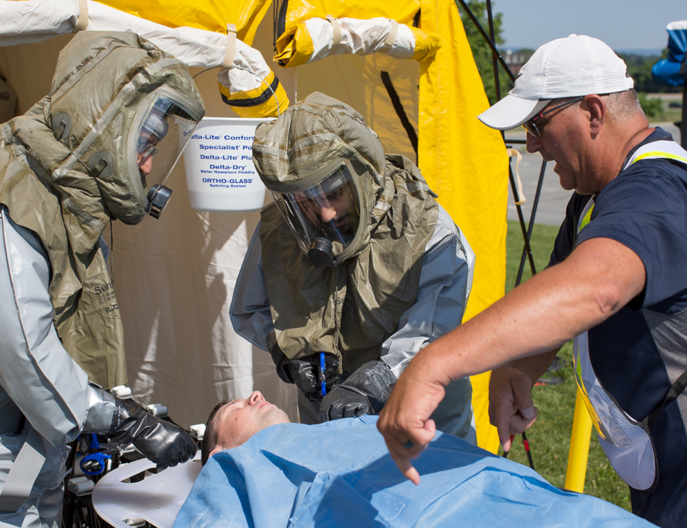 Two men in hazmat suits tend to a man laying on a stretcher. A third man, at right, wearing a short-sleeve shirt and ballcap, talks with the other men.