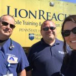 Three people “ two men and one woman “ pose for a photo in front of a large trailer that has the word LionReach on it as well as the Penn State Health Milton S. Hershey Medical Center logo.