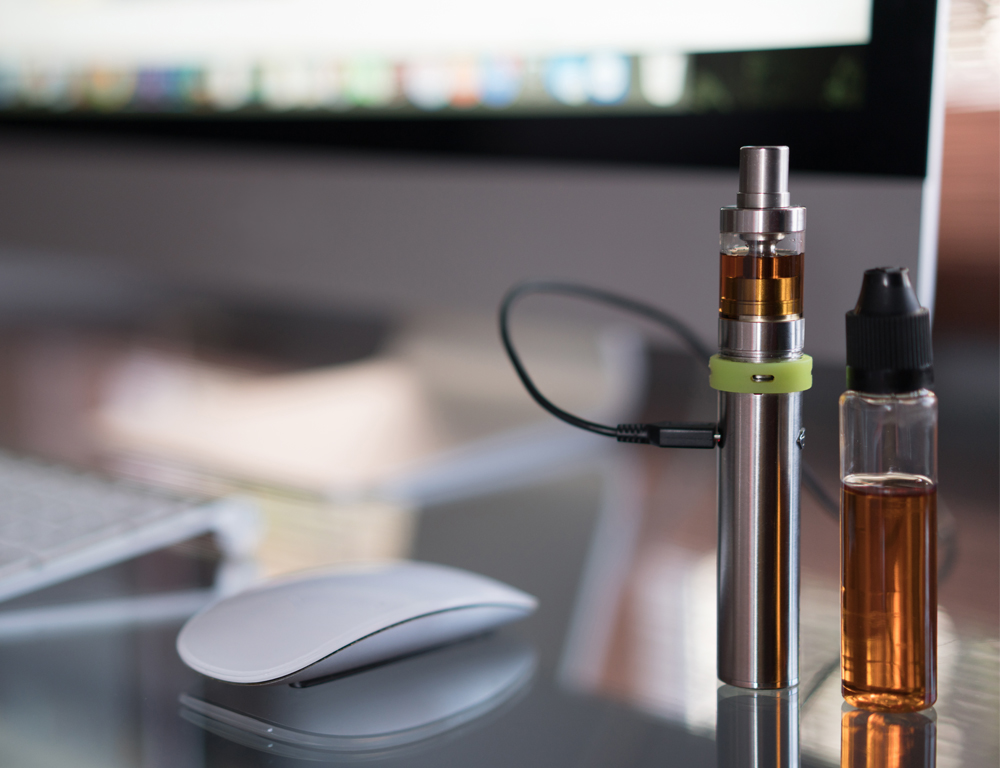 A close-up of a vaping device and a small vial of vaping liquid sitting on a counter. A computer keyboard, monitor and mouse sit nearby, slightly out of focus.