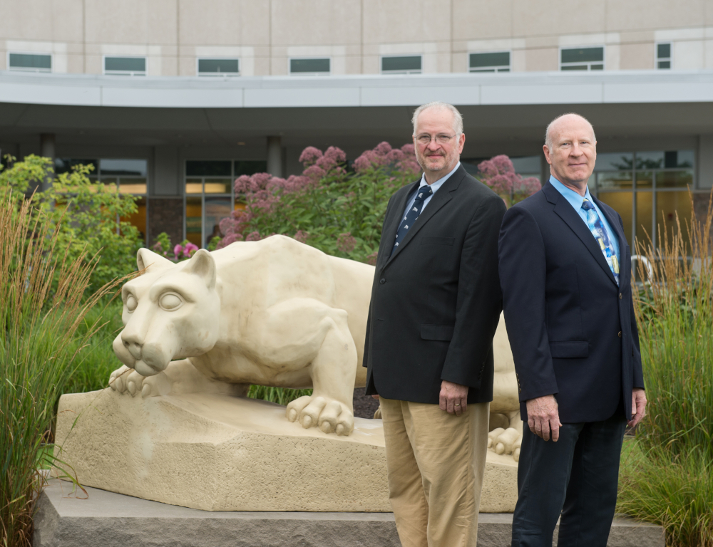 Two men in suits stand back-to-back, slightly off center to the right. A Penn State Nittany Lion statue is at left, and a building is in the background.