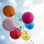 A set of eight balloons in different colors are seen against the background of a cloudy sky.