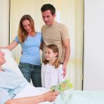 A stock photo shows a woman in a hospital bed with a family of three - a couple and a child - looking at her and smiling.