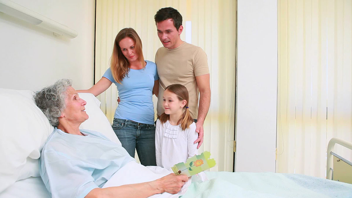 A stock photo shows a woman in a hospital bed with a family of three - a couple and a child - looking at her and smiling.