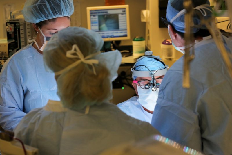 Four people in surgical scrubs and gowns are gathered around a patient in the operating room. A computer monitor and some equipment are visible in the background.