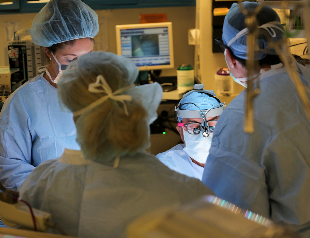 Four people in surgical scrubs and gowns are gathered around a patient in the operating room. A computer monitor and some equipment are visible in the background.