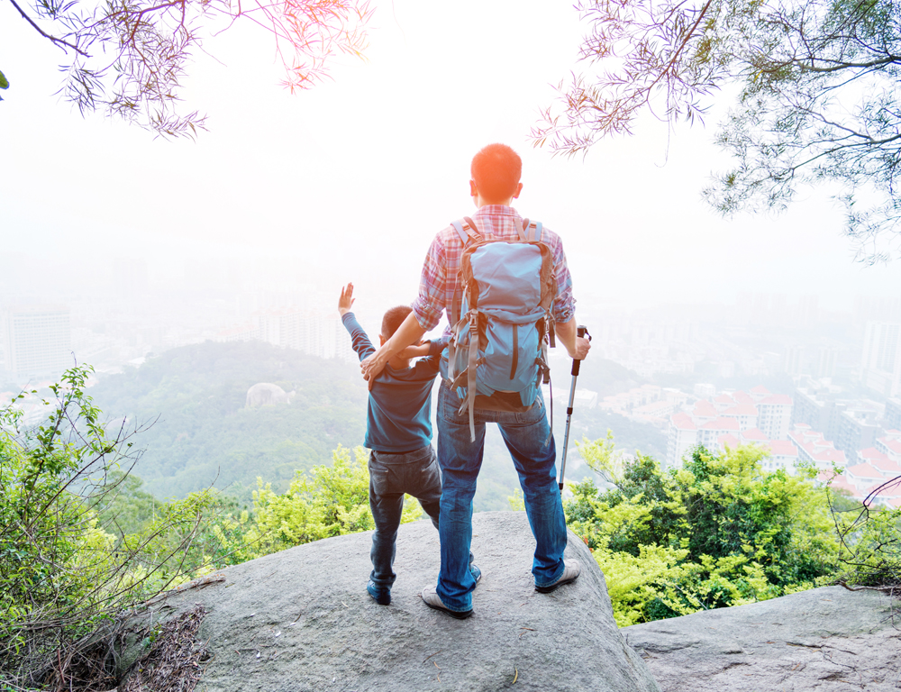 The view from behind as a man and child, dressed in hiking gear, stand on a rock looking off the edge of a mountain. Trees are in the foreground. Buildings and more trees and hills are in the background.