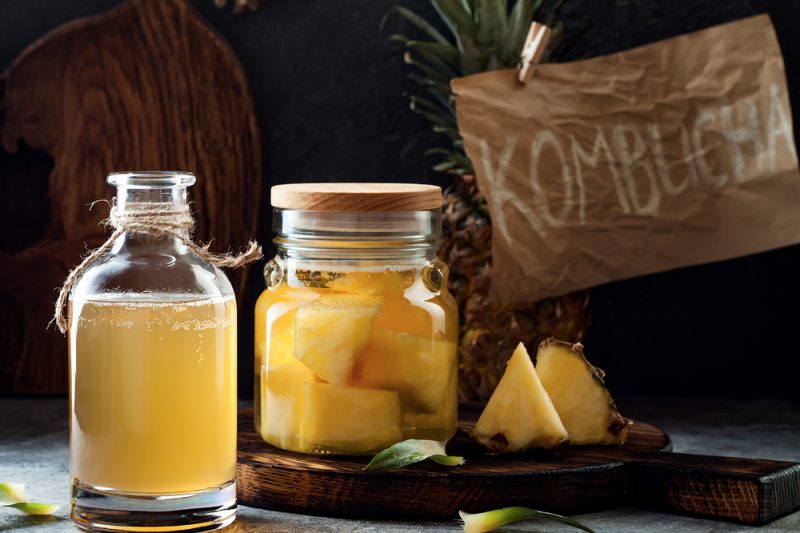 In the foreground are two glass jars with yellow liquid, one containing pieces of pineapple. In the background is a pineapple and a sign reading “KOMBUCHA.”
