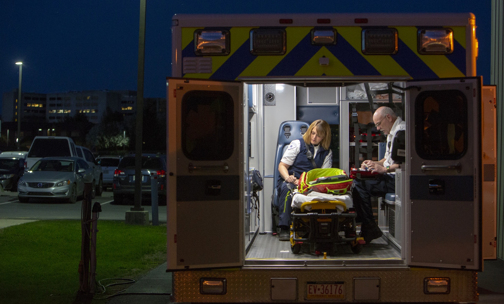 Amber Lewis, left, an EMT with Penn State Health Life Lion EMS, and paramedic Dave Heim sit in the back of a Life Lion ambulance at night. The doors of the ambulance are open, and the ambulance is in a parking lot. Amber is writing on a notepad, and Mike adds supplies to a medical kit. Another medical kit is lying on a gurney between them.