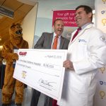 A man in a suit hands a giant check to a man in a medical coat. Both men are smiling as the Nittany Lion mascot looks on.