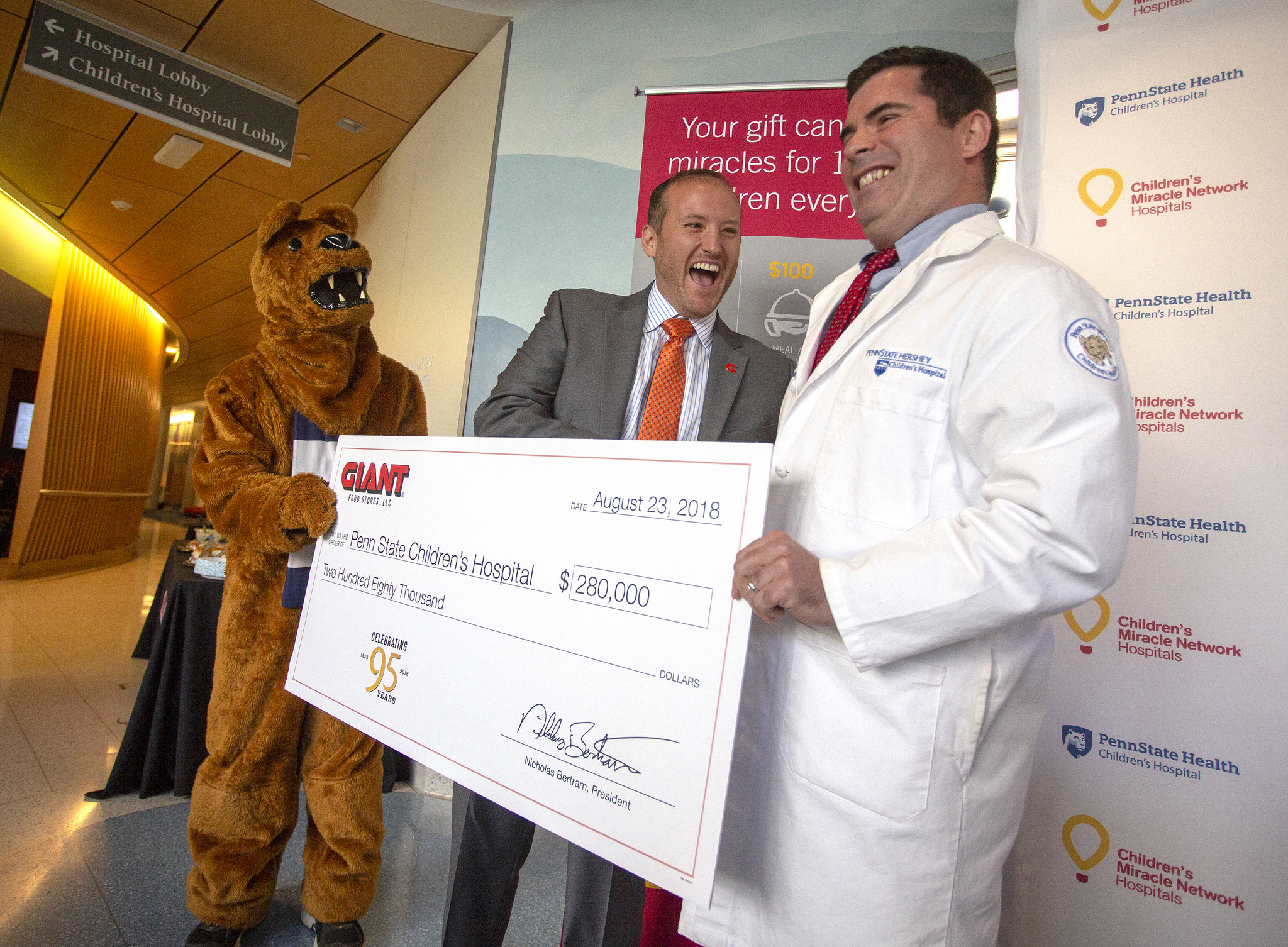 A man in a suit hands a giant check to a man in a medical coat. Both men are smiling as the Nittany Lion mascot looks on.