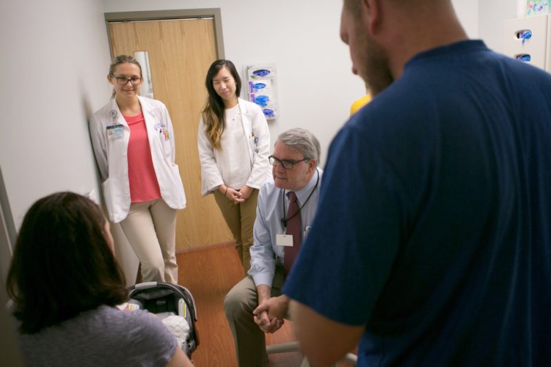 In a clinic room, a man in a tie speaks with a woman who’s holding an infant and facing away from the camera. Three other people are in the room, standing and looking on.