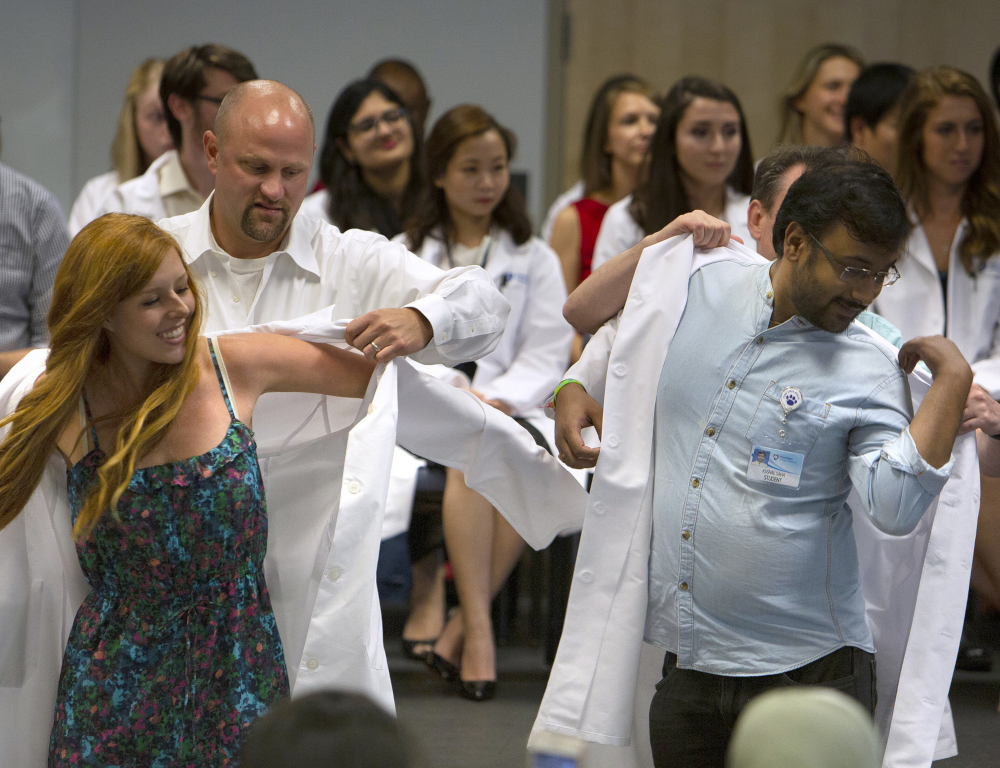 A young woman and a young man in the foreground are each getting help putting on white clinical/lab coats from people standing behind them. In the background, several people wearing white coats are seated, pictured slightly out of focus.