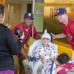 Two Harrisburg Senators baseball players stand on either side of a child in a mobile hospital bed while a woman standing to the side takes their picture.