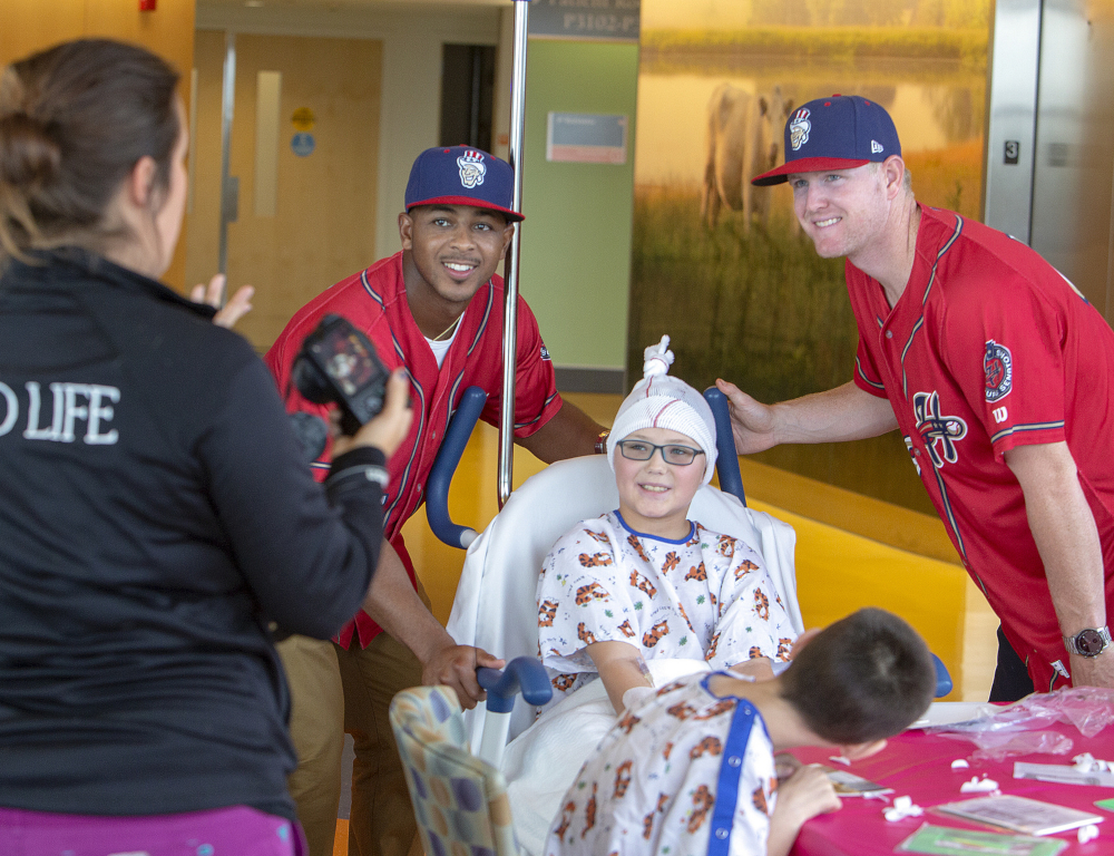 Two Harrisburg Senators baseball players stand on either side of a child in a mobile hospital bed while a woman standing to the side takes their picture.