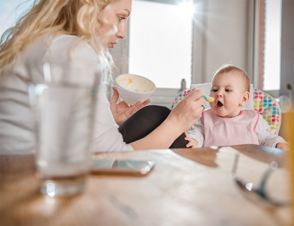 A woman sits next to her baby, who's seated in a high chair, and feeds her with a spoon. A baby bottle is in the foreground; a window is in the background.