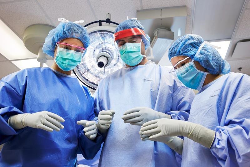 Three medical professionals dressed in operating room scrubs, caps and face masks stand in an operating room.