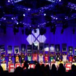 A group of young people stand on a stage holding up individual numbers that spell $7,036,560.82 and letters that spell out FOR THE KIDS.