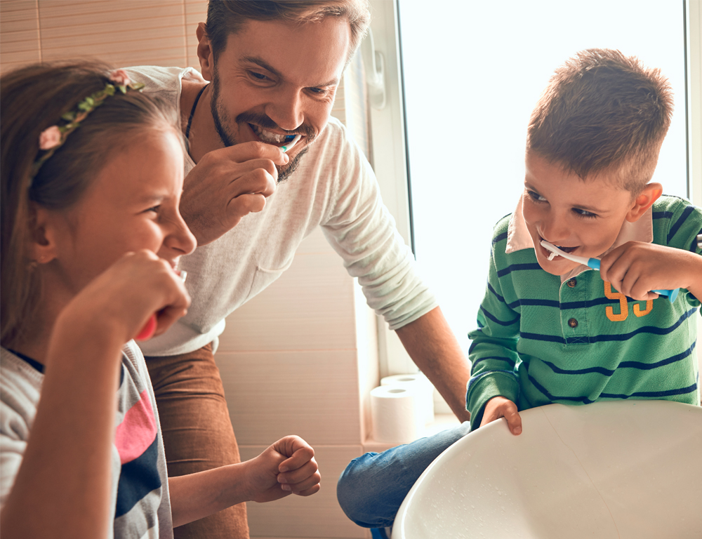 Two young children, a boy and a girl, stand with their father in front of a bathroom sink. All three of them are brushing their teeth.