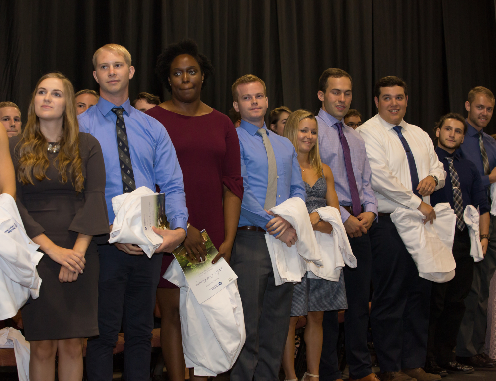 Several medical students stand in a row, holding in front of them their white medical coats.