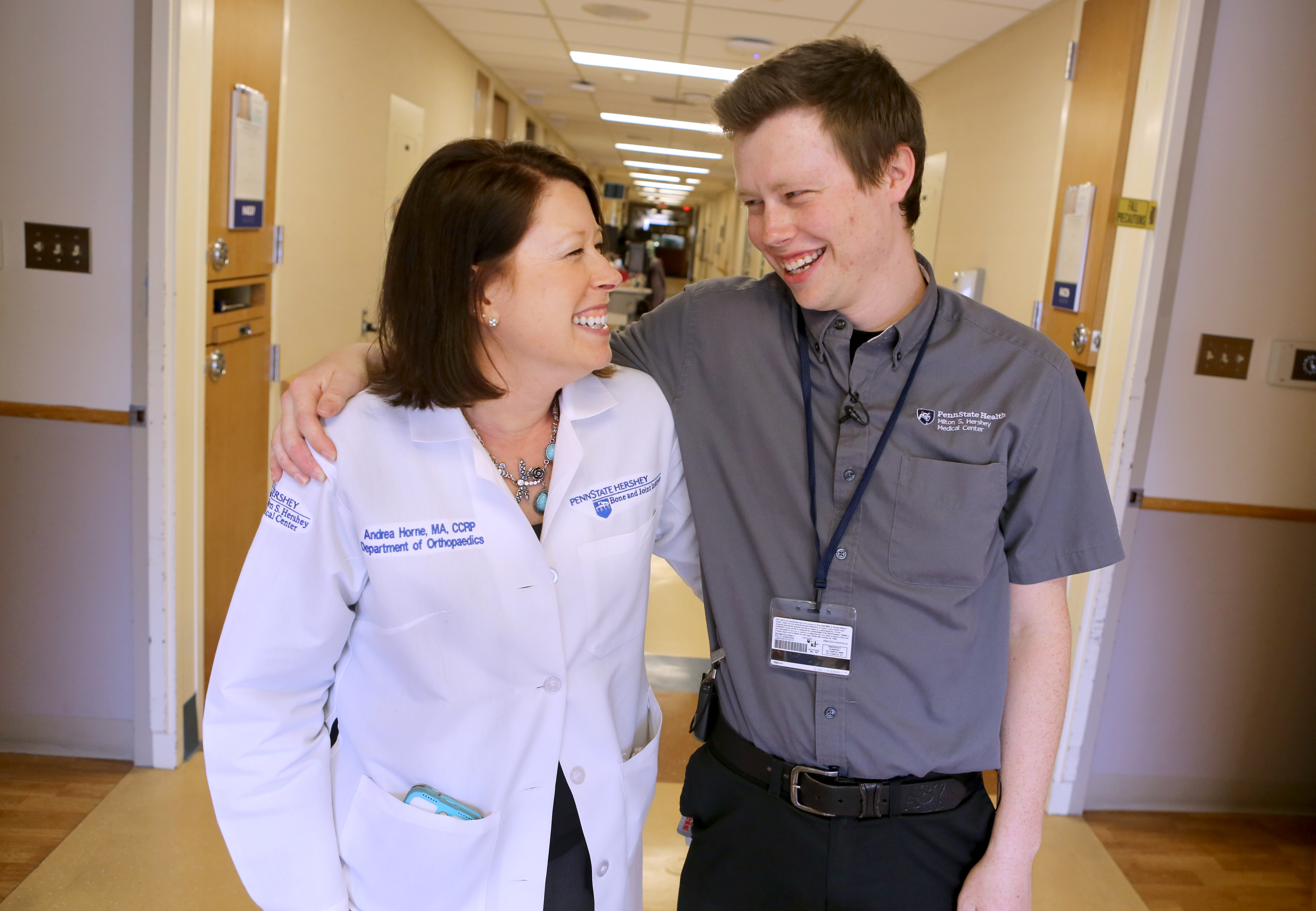 Andrea and Cole Horne stand in a hallway at Hershey Medical Center with their arms around each other and smiling broadly. Andrea is wearing a white lab coat with a Department of Orthopedics logo. Cole is wearing a button-down shirt with a Hershey Medical Center logo.