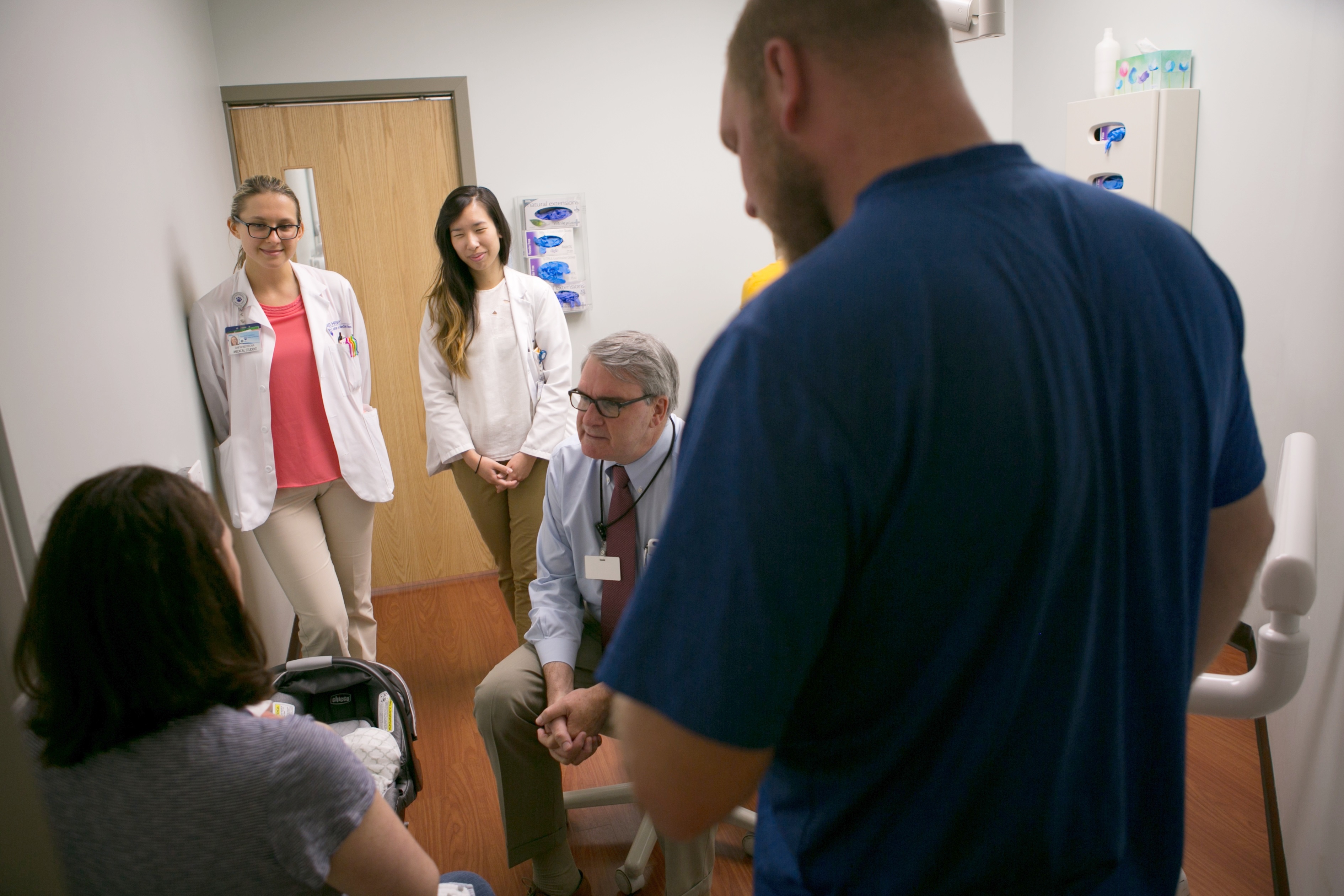 Dr. Donald Mackay sits on a stool and and talks to two parents in a doctor’s office. Their baby’s car seat is on the floor. Two female clinicians stand next to the doctor smiling.