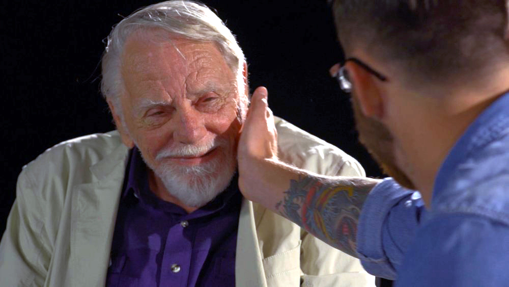 Caleb and David are two participants in the FaceAge project, where younger and older participants are seen studying and describing one another’s faces. An older man with a beard is pictured facing the camera, and a younger man's head is visible from behind; the younger man is touching the face of the older man.