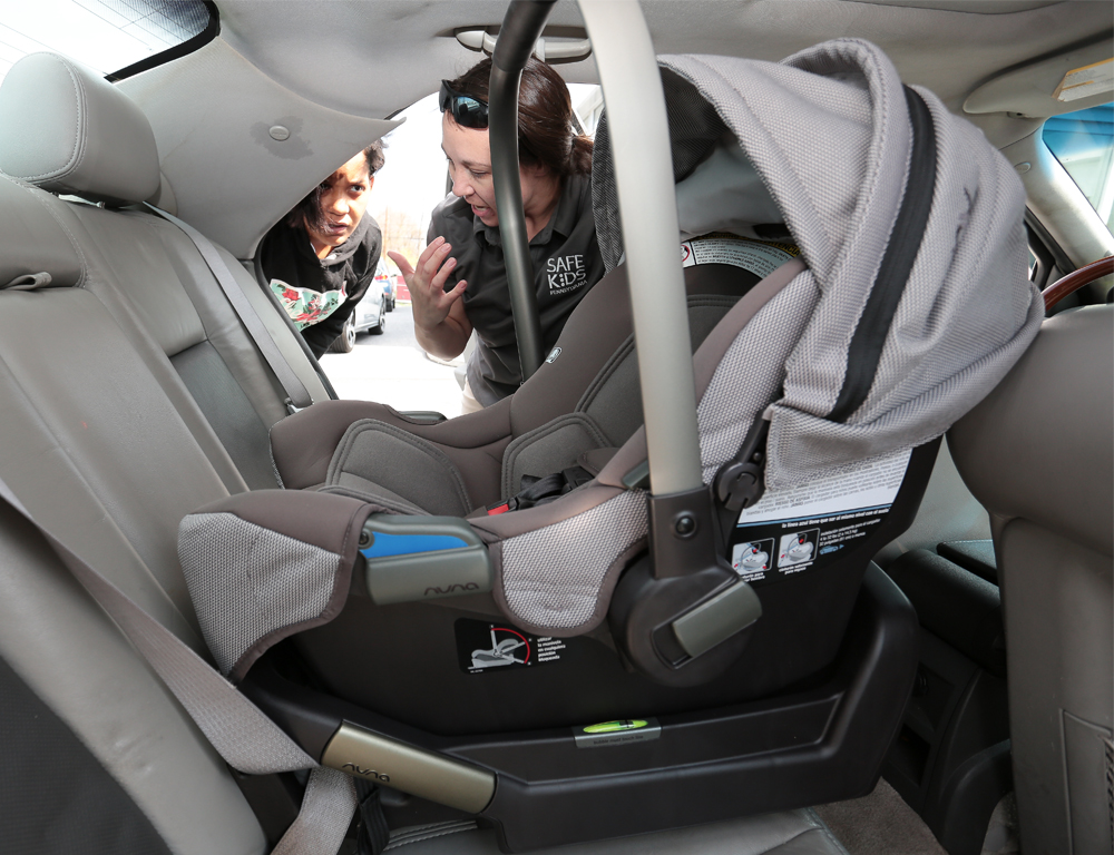 The view from the back seat of a car, across a rear-facing infant car seat, as two women peer in through the opposite side door, talking.