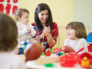 Photo focuses on a woman who is smiling and clapping her hands. She is seated at a table with various toys on top of it with two young children. Blurred out in the foreground is the back of another young child.