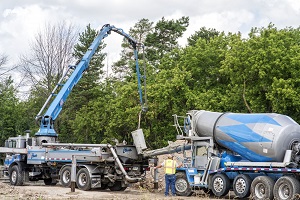 Two concrete pouring trucks outdoors. Trees are in the background. There is a workman in the foreground in front of one of the trucks with his back to the camera.