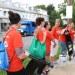 Six women stand along a sidewalk near a city street. Some hold bags; two are holding step ladders. They all wear orange shirts with the slogan “LIVE UNITED” and the United Way logo.