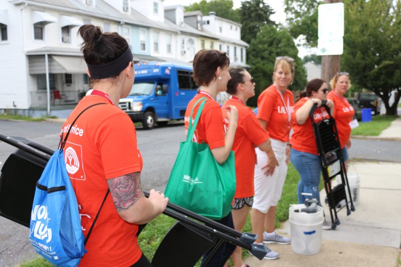 Six women stand along a sidewalk near a city street. Some hold bags; two are holding step ladders. They all wear orange shirts with the slogan “LIVE UNITED” and the United Way logo.