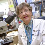 Dr. Elaine Eyster poses for a photograph in a lab, wearing a lab coat. A microscope is nearby.