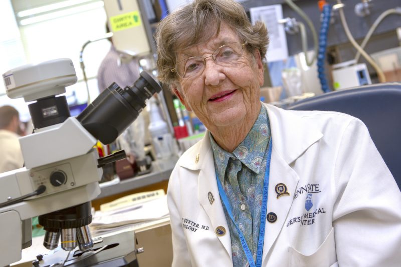 Dr. Elaine Eyster poses for a photograph in a lab, wearing a lab coat. A microscope is nearby.