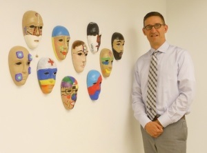 A man is pictured from the knees up standing next 10 masks with different designs hanging on a wall. He is wearing slacks, button down shirt, tie and glasses.