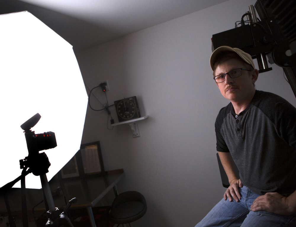A man wearing a ballcap, shirt and jeans sits on a stool in a photo studio. A camera and light panel are on the left side of the photo.